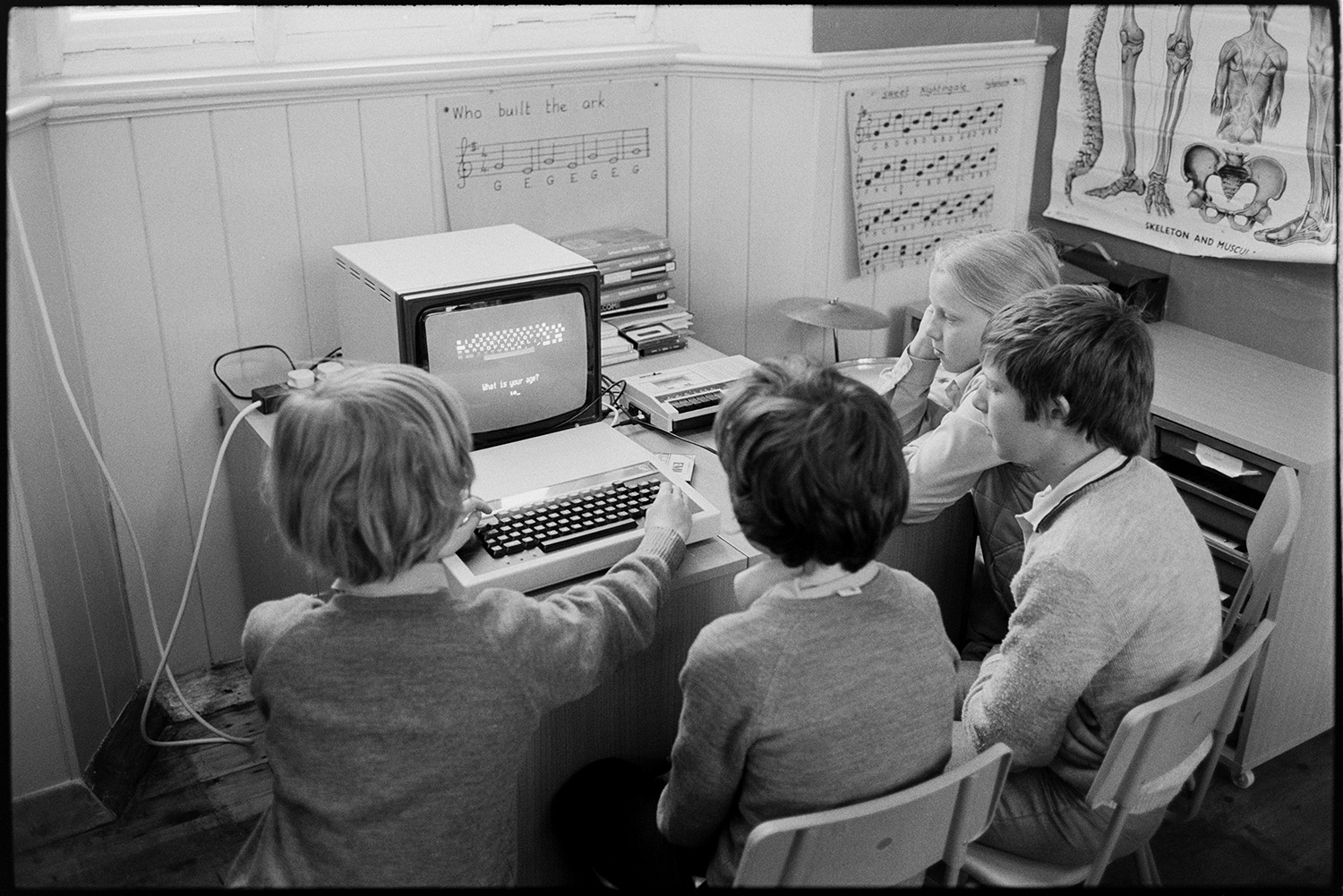 Schoolchildren using computer BBC Micro.
[Four children grouped round a BBC Micro computer in Dolton Primary School. Music and a poster of a human skeleton is displayed on the walls behind them.]