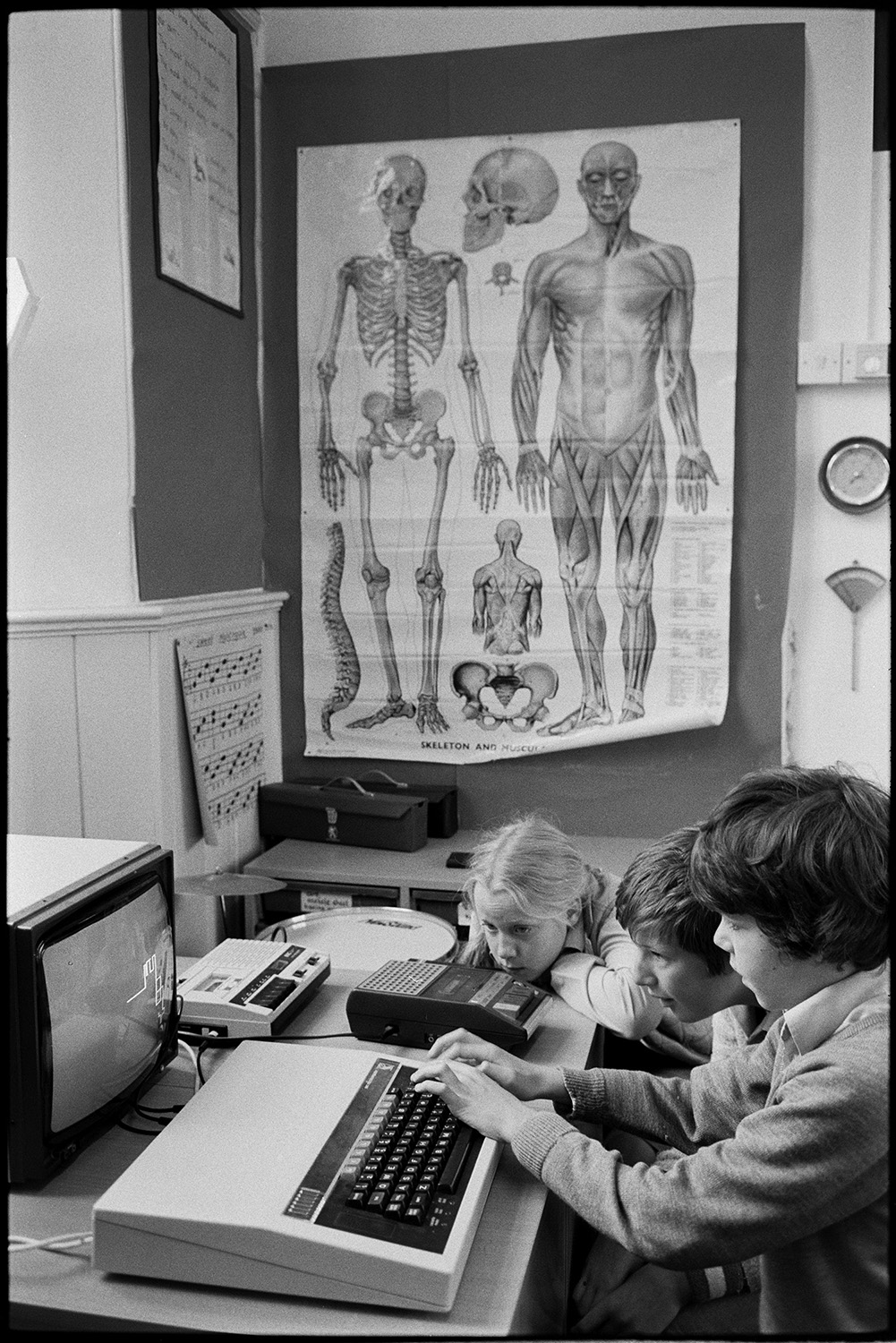 Schoolchildren using computer BBC Micro.
[Three children operating a BBC Micro computer at Dolton Primary School with a large poster of a human skeleton mounted on the wall behind them.]