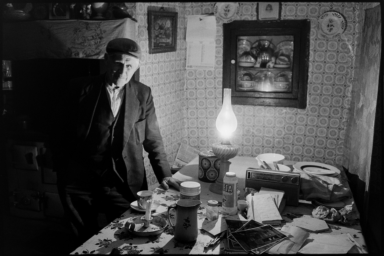 Farmhouse interior at night with oil lamp as light, farmer pouring tea, built in cupboard.
[Wilfie Spiers standing inside a room in his farmhouse at Mount Pleasant, Beamsworthy. he is stood by a table with a lit oil lamp, unlit candle in a holder, tea cup and saucer, jug, plates, radio, photographs and papers. A cupboard is built into the wall behind him containing china. A picture, a calendar and three china plates are hung on the wall which is covered with patterned wallpaper.]