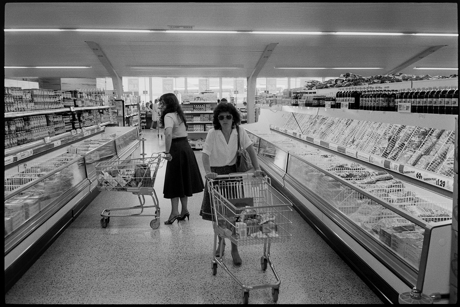 Interior of supermarket, people shopping, car park. Fruit, cash desk, check out.
[Two women pushing trolleys containing their shopping between two long chiller cabinets in Presto Food Market in Bideford. Bottles and cans are stacked on shelves above the freezer cabinets. One of the women is wearing a pair of sunglasses.]