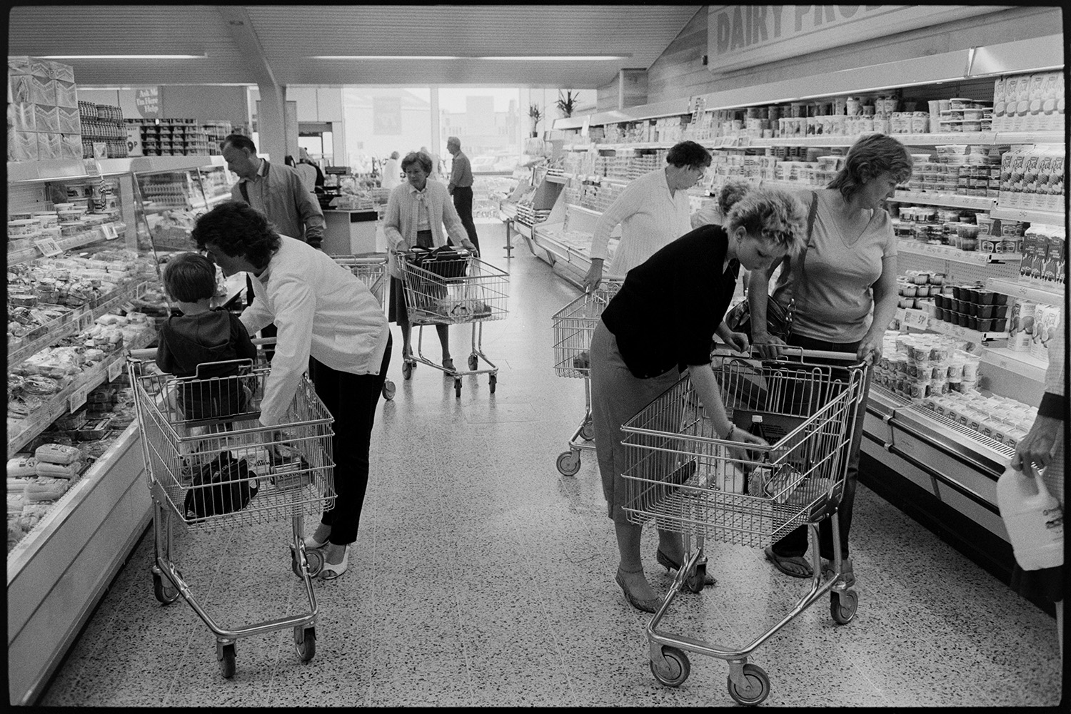 Interior of supermarket, people shopping, car park. Fruit, cash desk, check out.
[Women, men and children shopping in Presto Food Market in Bideford. They are placing items in trolleys in an aisle with chiller cabinets and shelves.]