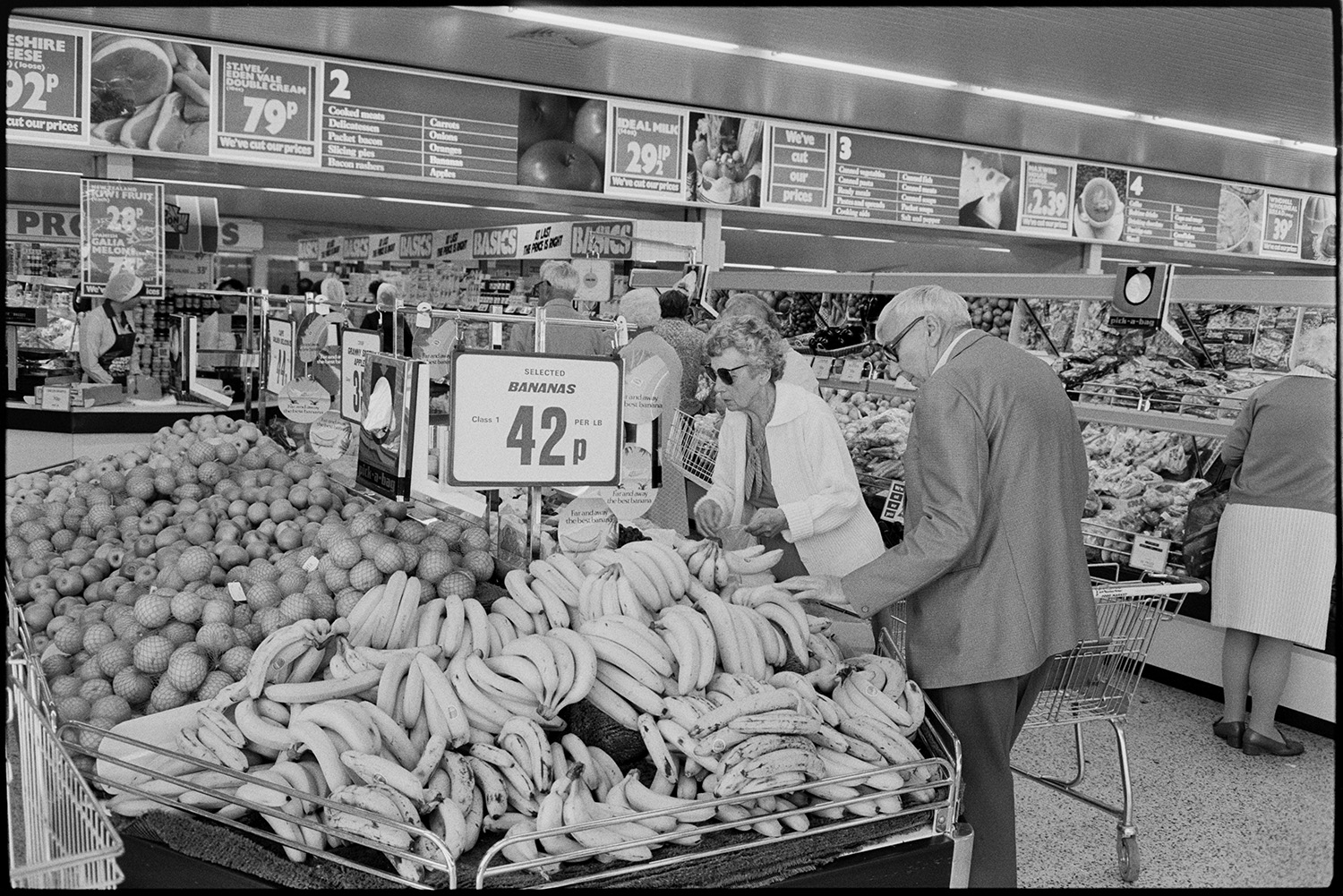 Interior of supermarket, people shopping, car park. Fruit, cash desk, check out.
[A man and a woman selecting bananas in Presto Food Market in Bideford and placing them in a shopping trolley. Other fruit is on display, including apples and oranges. There are a number of display boards showing details of products and prices.]
