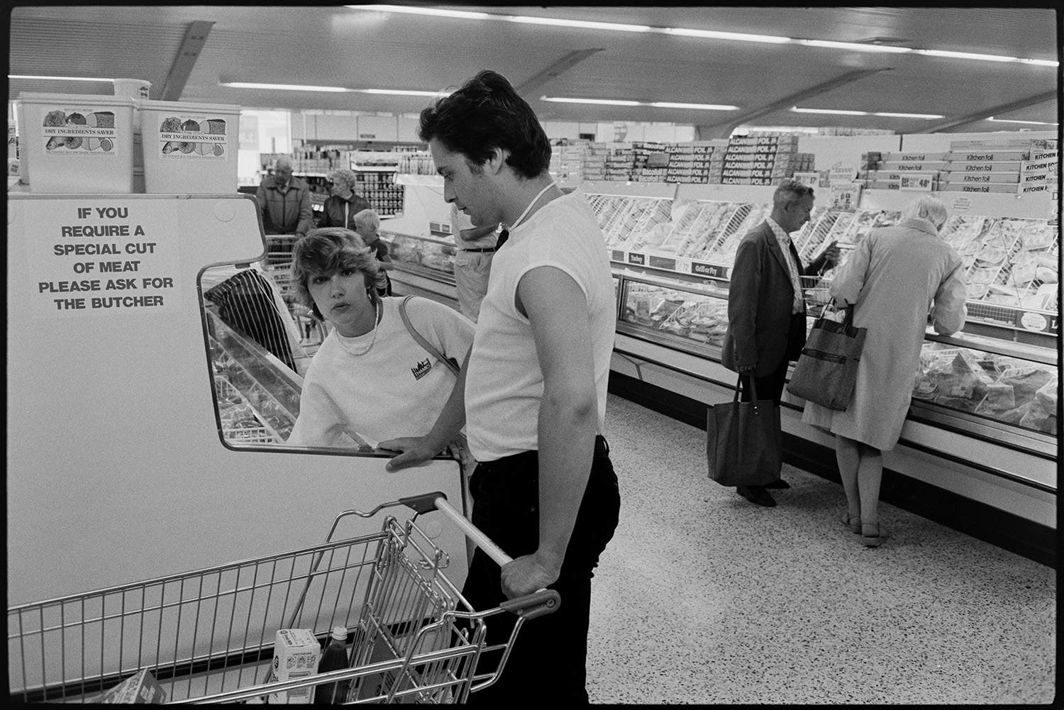 Interior of supermarket, people shopping, car park. Fruit, cash desk, check out.
[Women and men selecting items from chiller cabinets in the Presto Food Market in Bideford and placing the items in shopping trolleys. A sign on one of the cabinets reads 'If you require a special cut of meat please ask for the butcher'.]