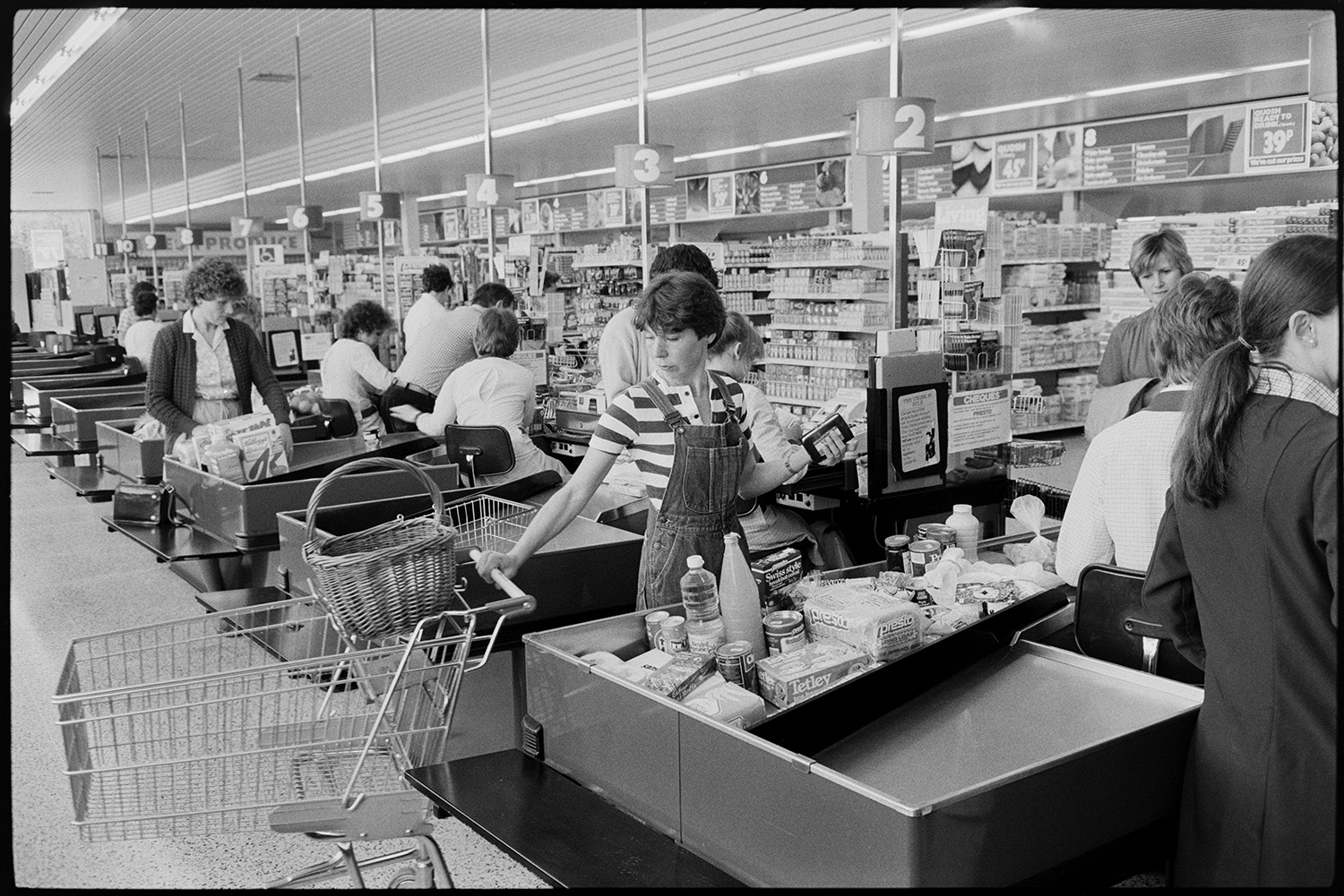 Interior of supermarket, people shopping, car park. Fruit, cash desk, check out.
[Customers at Presto Food Market in Bideford paying for their shopping at cash tills. One woman has a shopping trolley and a basket. Shelves stacked with good are visible in the background.]]