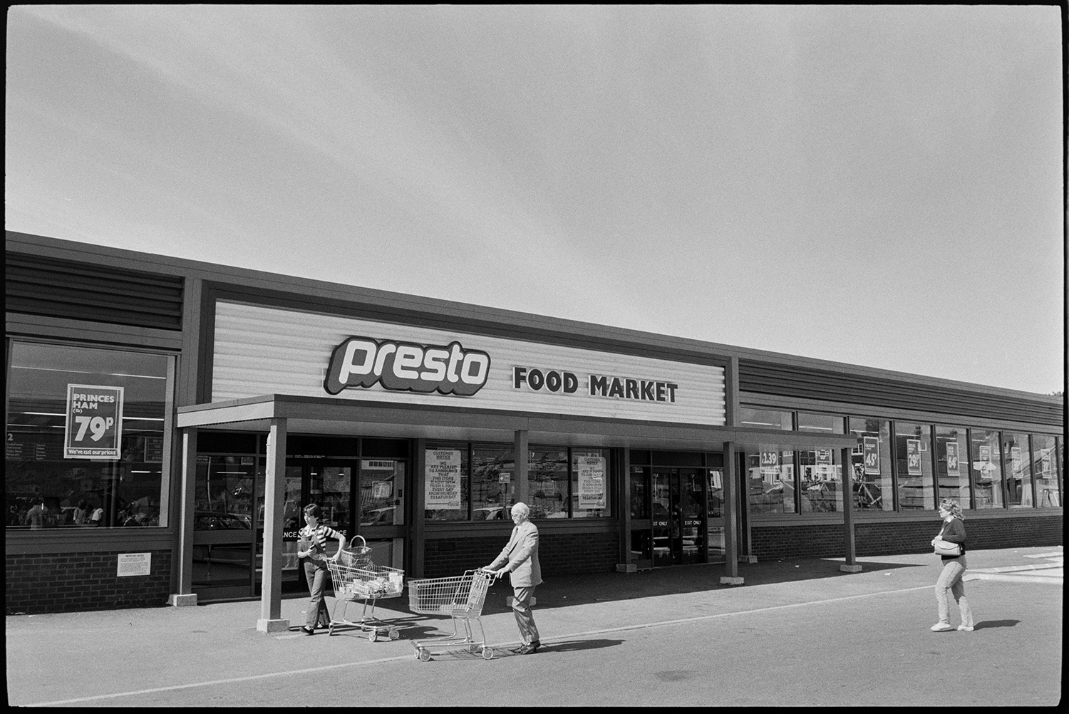 Interior of supermarket, people shopping, car park. Fruit, cash desk, check out.
[The entrance to Presto Food Market in Bideford, with three shoppers outside pushing shopping trolleys.]