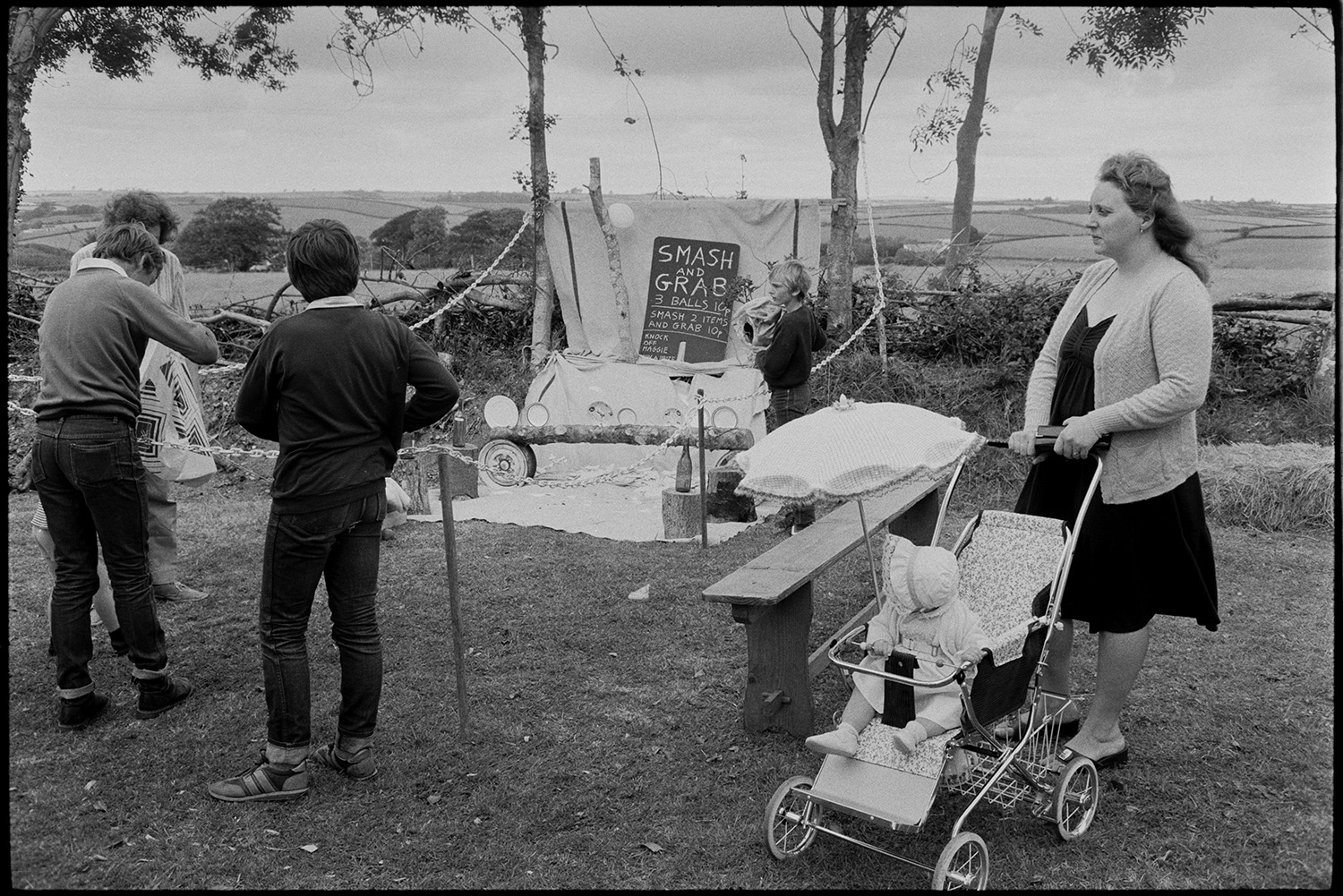 Garden fete, stalls, cakes, smash and grab.
[Man, woman and three children standing in front of a Smash and Grab game between two trees at a fete being held at Lanes End, Beaford. The woman is pushing a baby under a parasol in a pushchair. A landscape with trees, fields and hedgerows is visible in the background.]