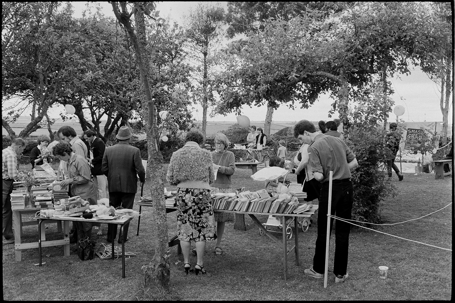 Garden fete, stalls, cakes, smash and grab.
[Men, women and children browsing bookstalls on tables under trees in the grounds of Lanes End, Beaford at a Garden Fete.]