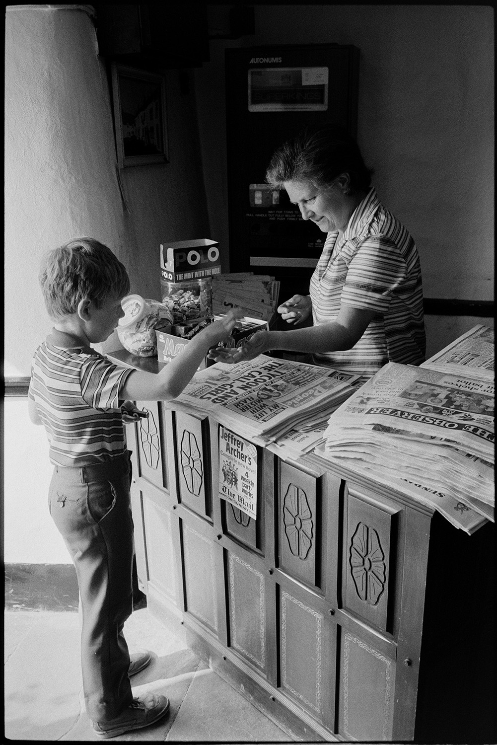 Sunday newspaper seller at door of pub, people buying papers.
[A boy buying sweets from a woman behind a carved wooden counter in the Rams Head Inn, Dolton on which there are sweets and piles of Sunday papers for sale. On the front of the counter is an advertisement for Jeffrey Archer's new novel to be published in four parts in The Mail on Sunday.]