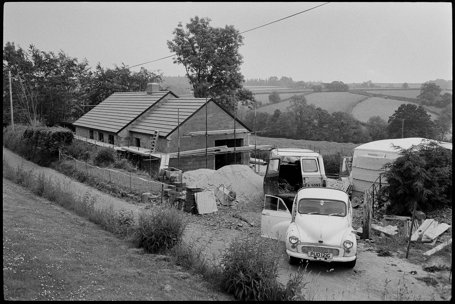 New house under construction.
[Two vans and a caravan parked beside a house being constructed in Aller Road, Dolton. Scaffolding is on the house. A landscape of fields and trees is visible in the background.]