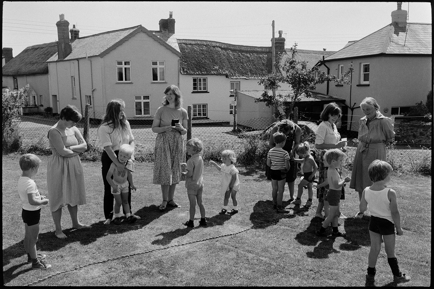 Sports day, children's races, parents egg and spoon, mothers women chatting, refreshments.
[Catherine Simmons, stood in the centre and possibly holding a wallet, talking with five other women and nine children at the start of an event at Dolton Sports Day. Thatched houses are visible in the background.]