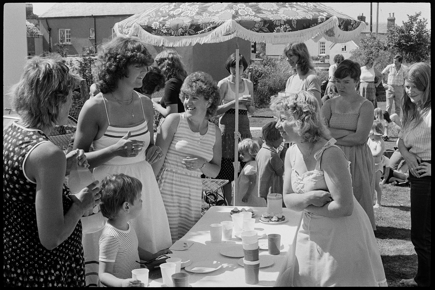 Sports day, women mothers with babies, children chatting and serving refreshments.
[A group of women and children chatting around a parasol covering a refreshment table at Dolton Sports Day.]