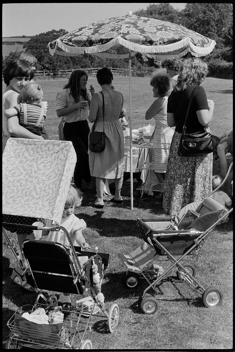 Sports day, women mothers with babies, children chatting and serving refreshments.
[Women chatting under a parasol covering a refreshment table at Dolton Sports Day, with pushchairs and children in the foreground.]