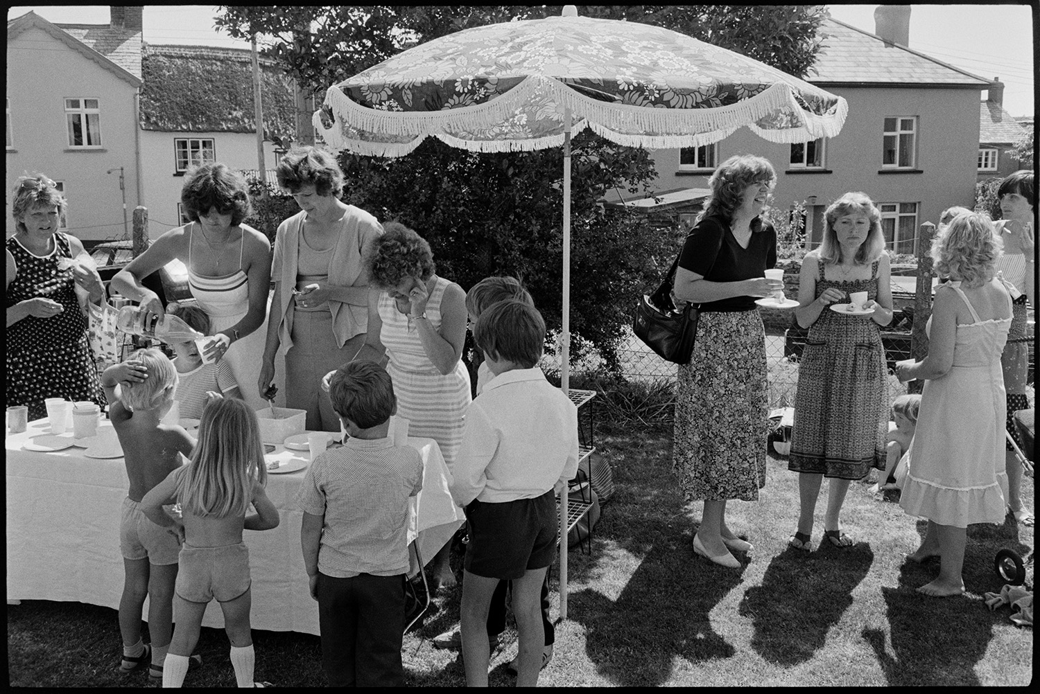 Sports day, women mothers with babies, children chatting and serving refreshments.
[Women serving drinks at a refreshment table to children and chatting under a parasol at Dolton Sports Day.]