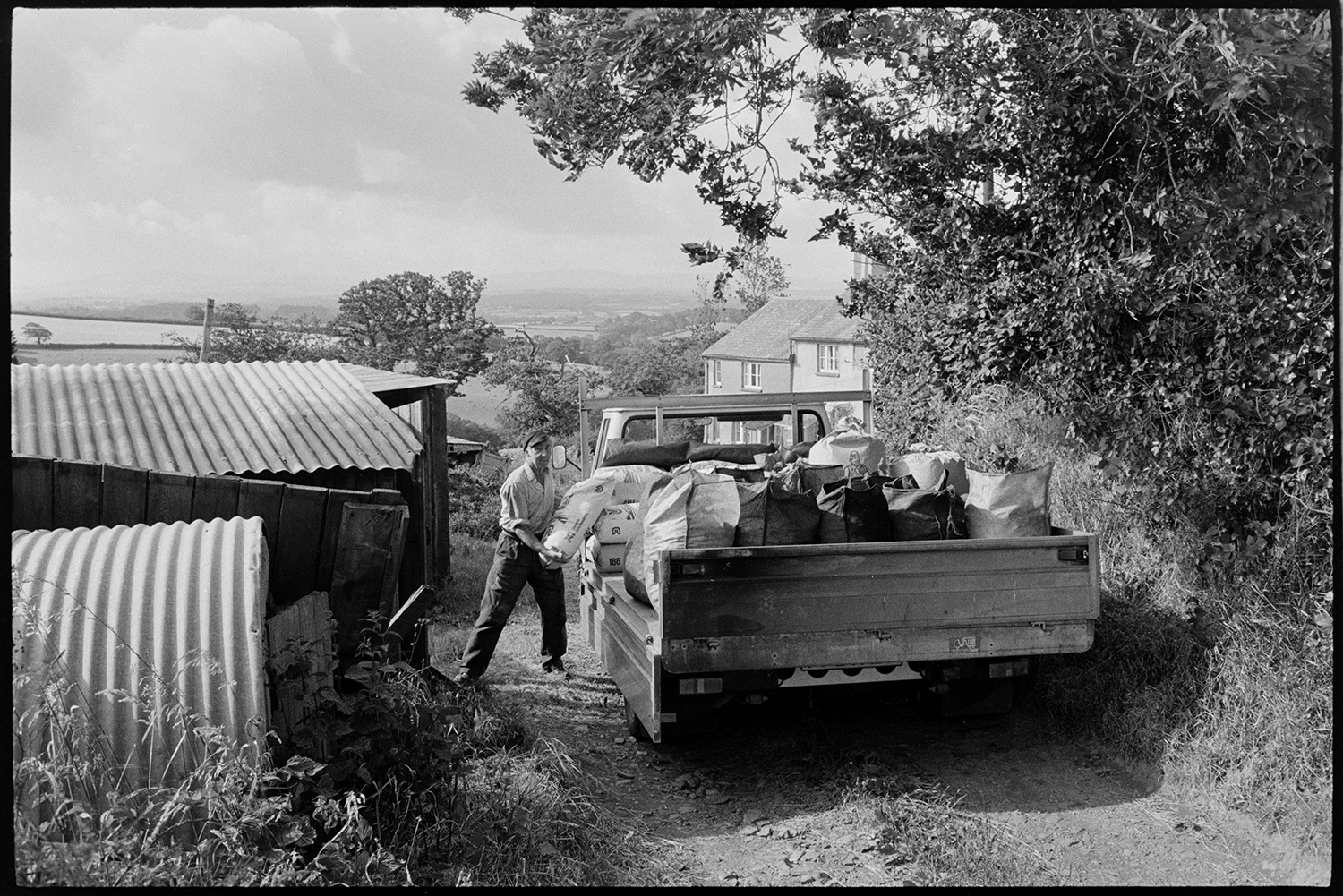 Man delivering, unloading sacks of coal from lorry.
[Mr Nott unloading sacks of coal from a truck parked at the entrance to Berry Farm, Iddesleigh, next to corrugated iron sheds. The farmhouse and a view of surrounding countryside can be seen in the background.]