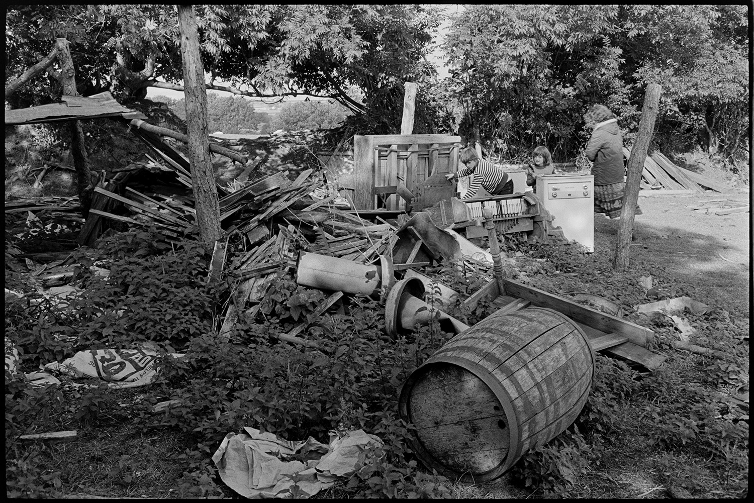Junk in corner of field.
[Two children and a woman looking at a pile of overgrown junk in the corner of a field at Ashwell, Dolton, including a wooden barrel, a washing machine and timber.]