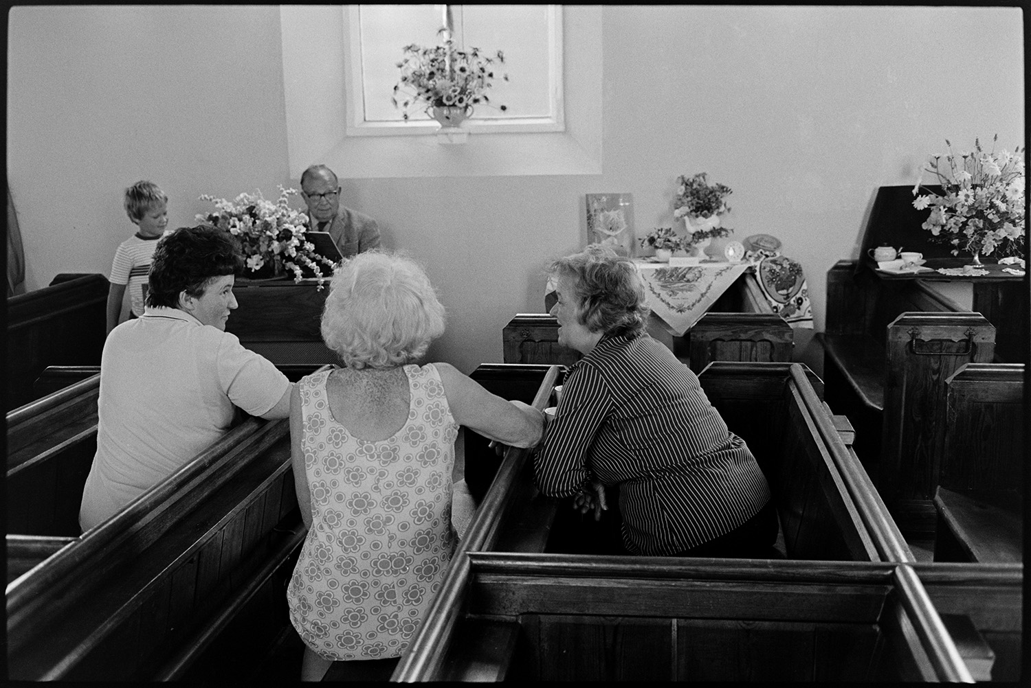 Baptist Chapel flower festival Minister playing organ, harmonium, mugs and biscuits, teas.
[Three women sitting in pews chatting at the Flower Festival in Dolton Baptist Chapel, with floral displays in the background. Minister Oliver is playing the organ or harmonium and a boy is turning the pages of the music for him.]