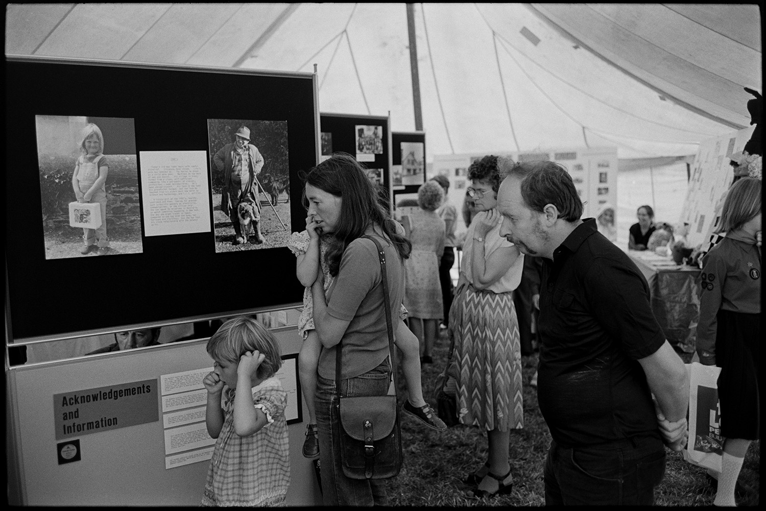 North Devon Show, Prize cattle, sheep, exhibitions etc.
[Interior of a marquee at the North Devon Show, near Alverdiscott. Visitors, including men, women and children, are looking at display boards showing photographs and information by James Ravilious. A woman is carrying a small girl in the foreground.]