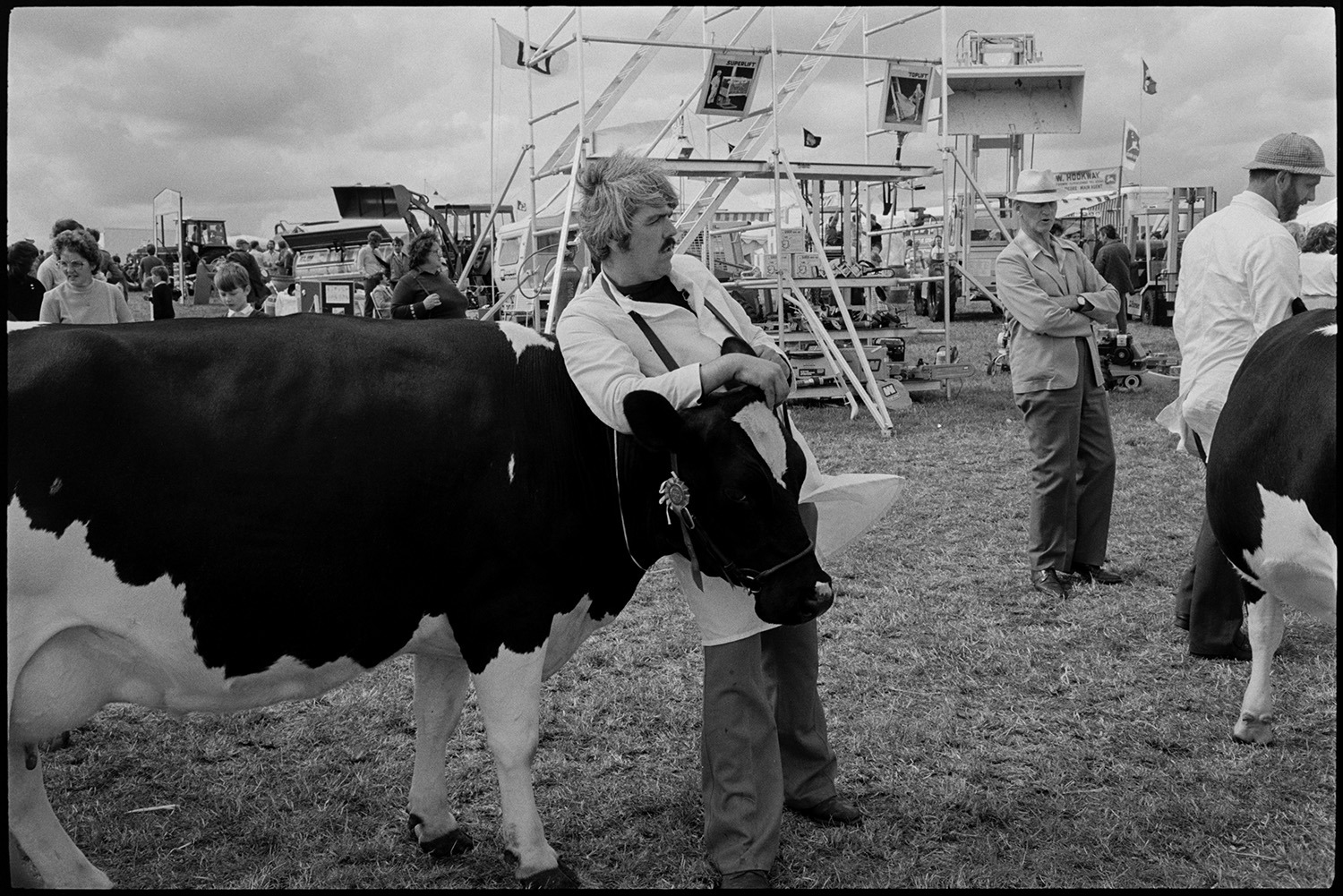 North Devon Show, Prize cattle sheep, people having teas, woman, mother and baby.
[Man with prize Friesian cow, at the North Devon Show near Alverdiscott. The cow is wearing a rosette. Machinery, including diggers and tractors, and visitors are visible in the background.]