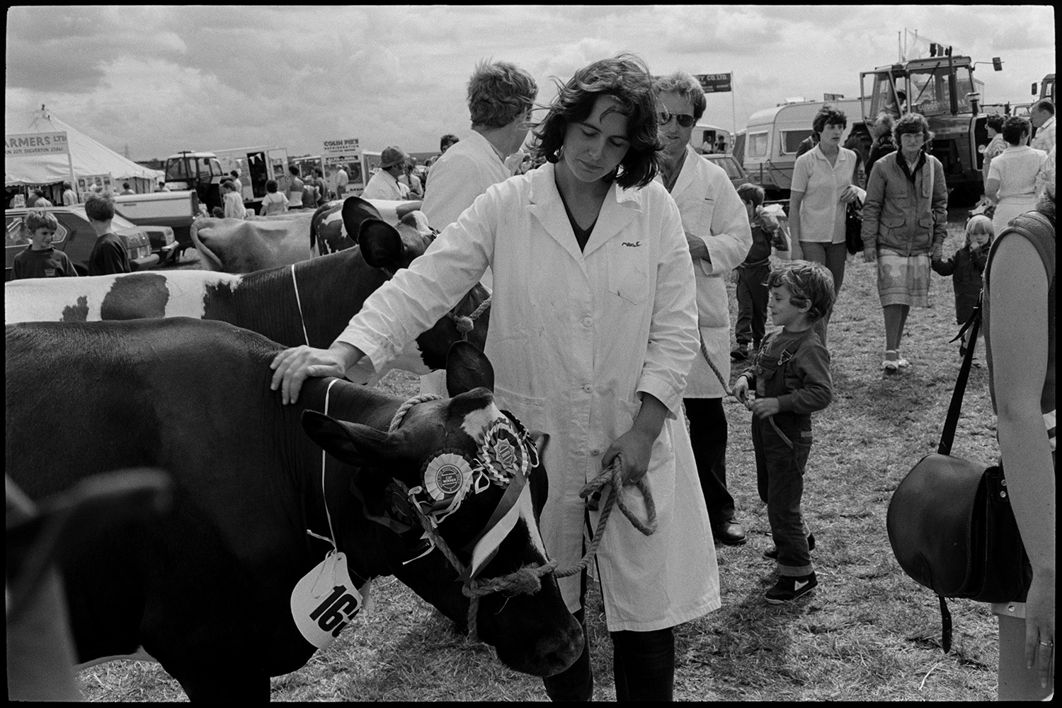 North Devon Show, Prize cattle, sheep, people having teas, woman, mother and baby.
[Woman exhibiting prize Friesian cow at the North Devon Show near Alverdiscott. The cow is wearing rosettes. Other exhibitors, visitors and stands are visible in the background.]