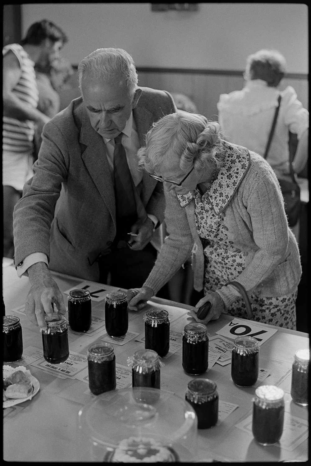 Flower show exhibits, jams, cups, sports, hot dog stall, bicycles, Clay Pigeon Shoot.
[Cyril Webber and a woman checking or judging the jam entries at the Dolton Flower Show in Dolton Village Hall.]