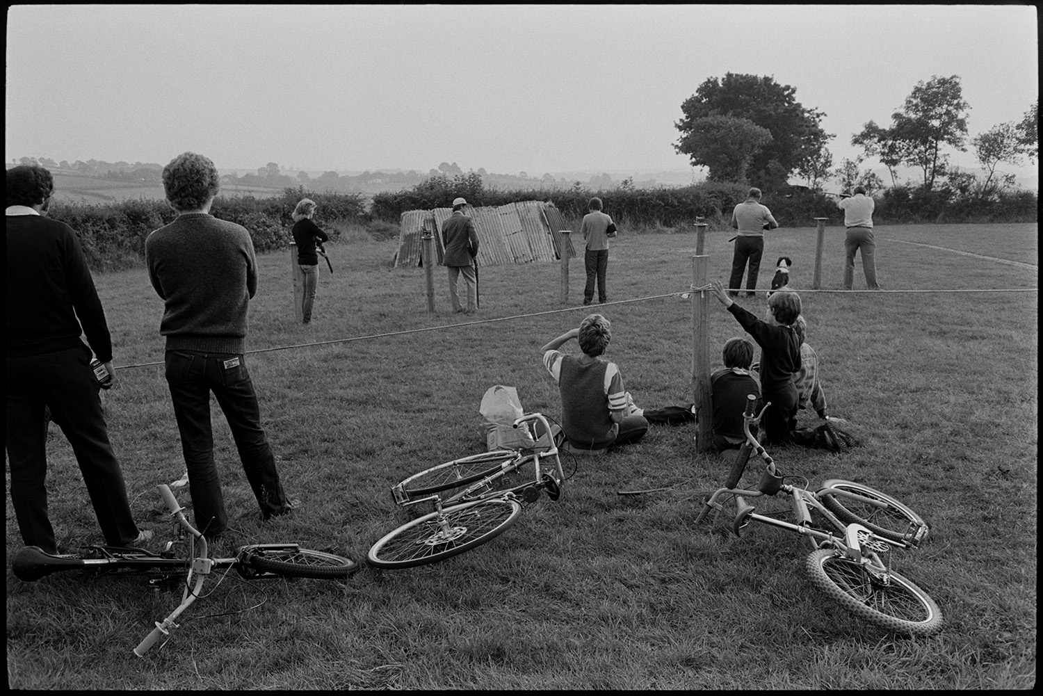 Flower show exhibits, jams, cups, sports, hot dog stall, bicycles, Clay Pigeon Shoot.
[Participants at the clay pigeon shoot at Dolton Flower Show. A group of boys are watching them behind a rope with bicycles lying on the ground.]