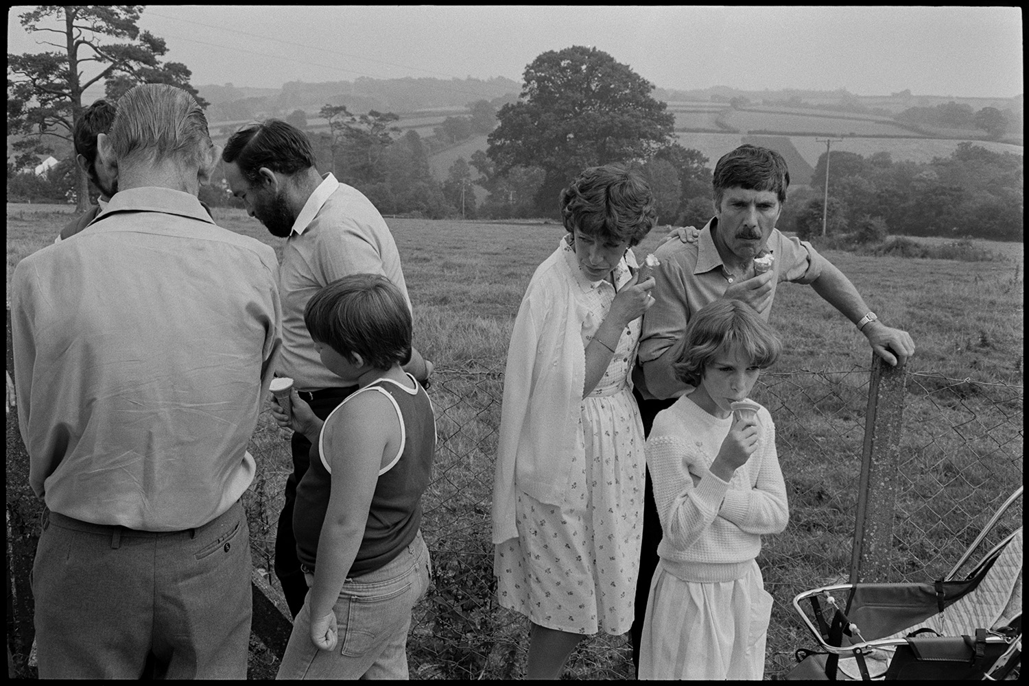 Flower show exhibits, jams, cups, sports, hot dog stall, bicycles, Clay Pigeon Shoot.
[Men, women and children at Dolton Flower Show eating ice creams, in a field with a wooded landscape in the background.]