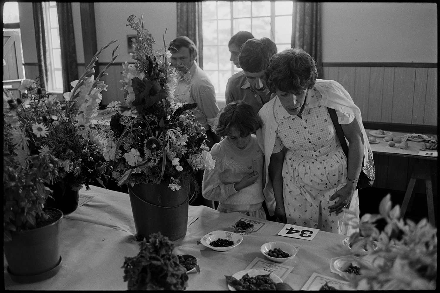 Flower show exhibits, jams, cups, sports, hot dog stall, bicycles, Clay Pigeon Shoot.
[A family at Dolton Flower Show looking at a table with a display of fruit and flower exhibits in Dolton Village Hall.]
