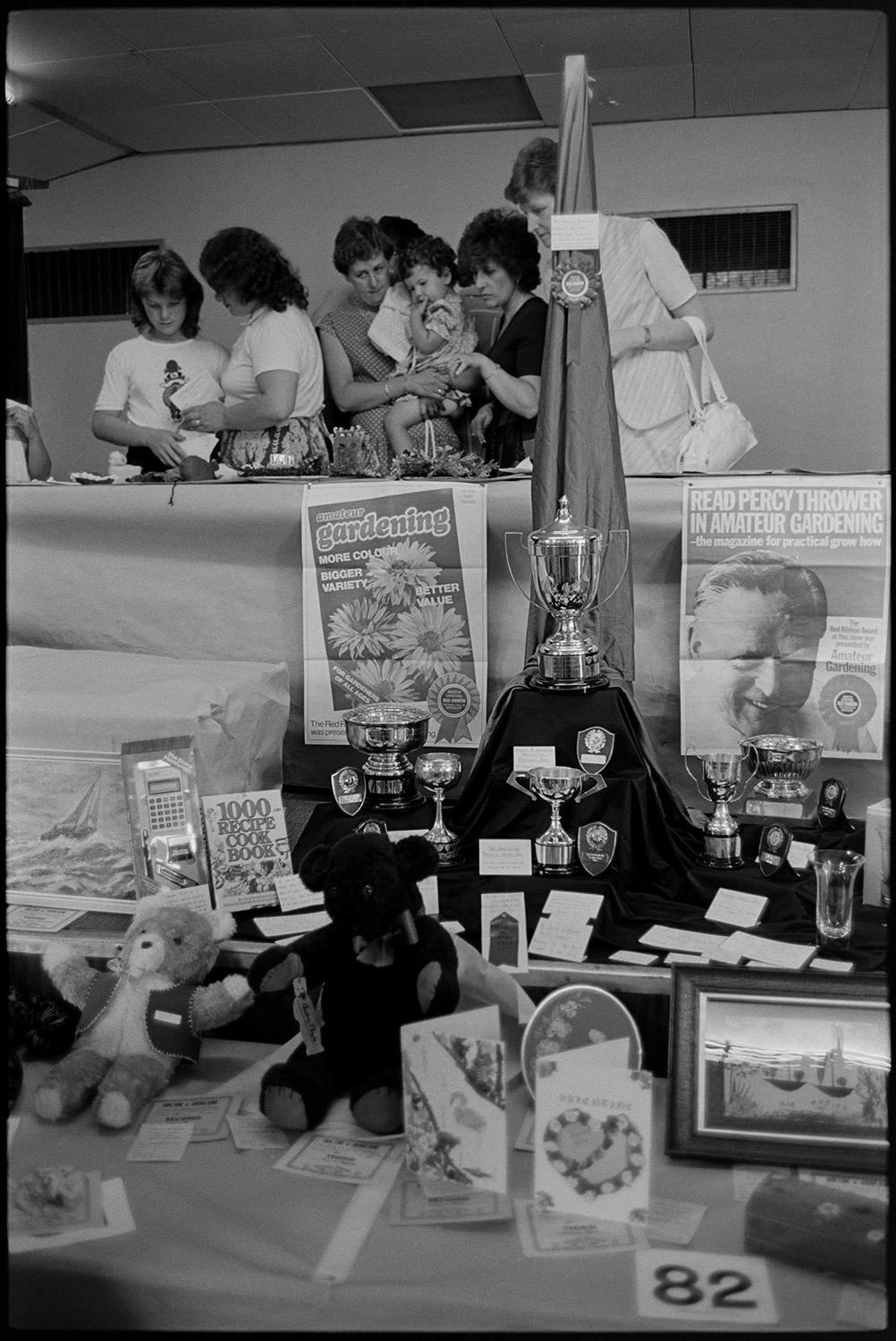 Flower show exhibits, jams, cups, sports, hot dog stall, bicycles, Clay Pigeon Shoot.
[Women and children at Dolton Flower Show, looking at exhibits on a table above a display of trophies, posters and prizes, in Dolton Village Hall.]