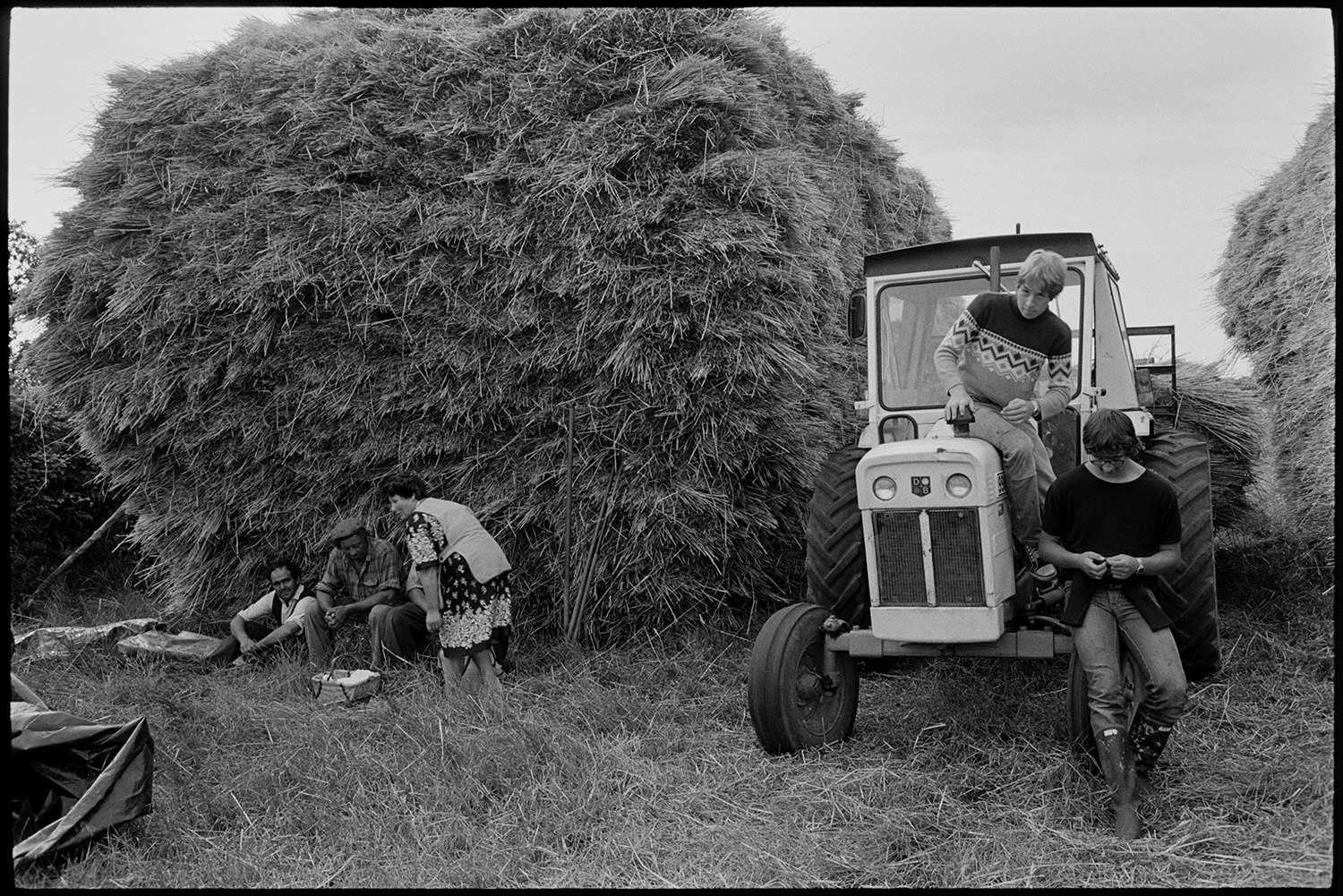 Men building wheat rick, having tea break, farmers wife with basket.
[Men building a wheat rick at Riddlecombe, having a tea break. Three men are sat under the rick and two other young men are leaning on a David Brown tractor nearby. A woman has brought them a basket of food.]