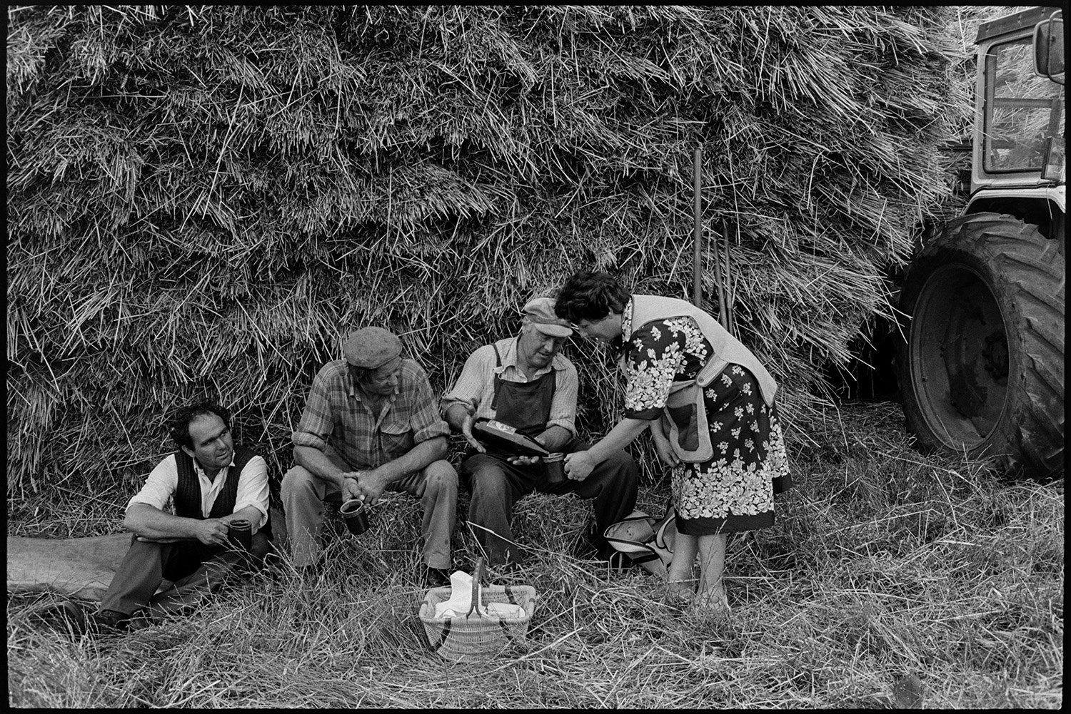 Men building wheatrick, having tea break, farmers wife with basket.
[Men having a tea break during building a wheat rick at Riddlecombe. They are sat under the rick. One of the men is pouring a drink from a bottle for the woman who has brought them a basket of food.]