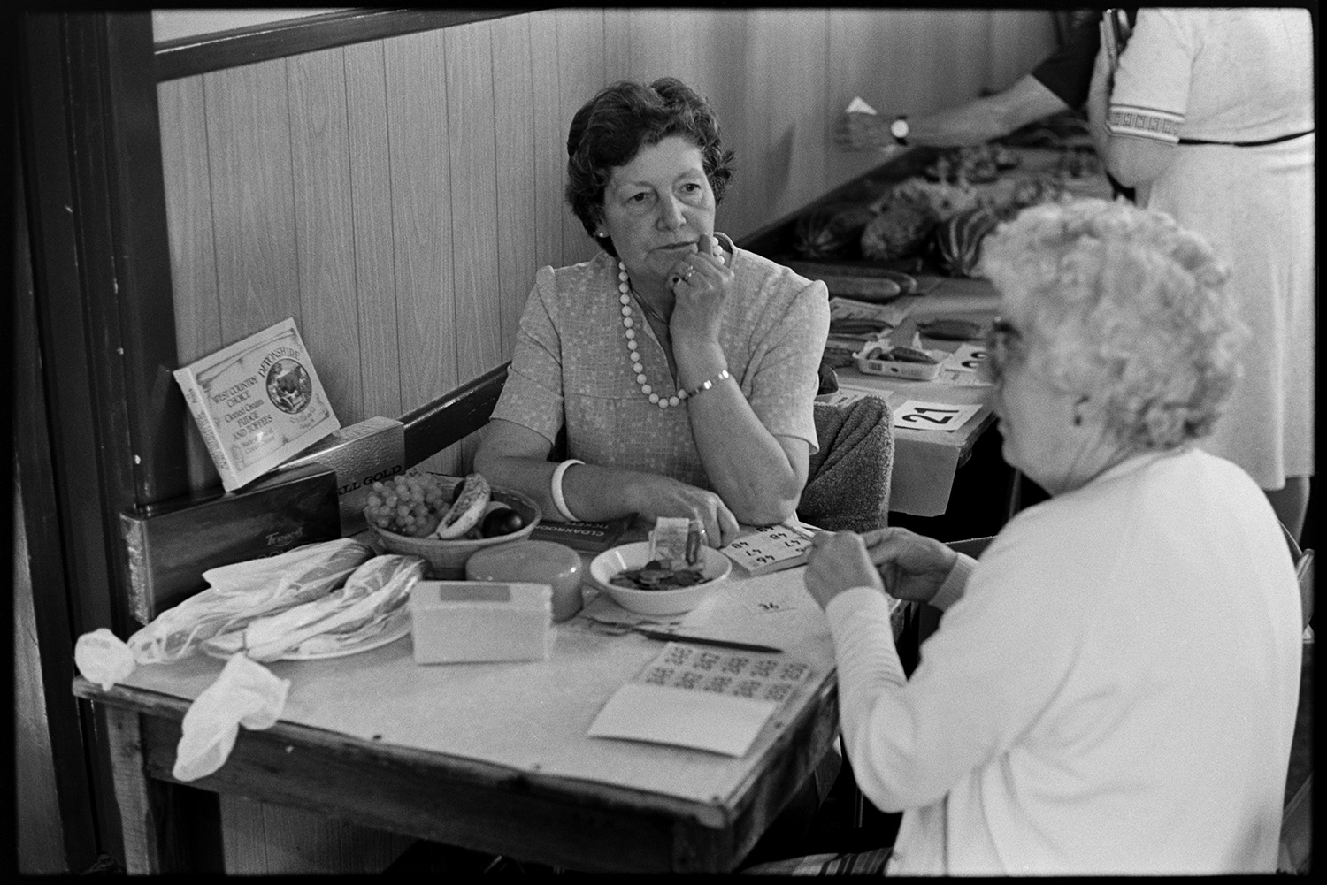 Flower show, people looking at exhibits, women looking at cake, paintings on display.
[Two ladies selling draw tickets at a table at the Dolton Flower Show in Dolton Village Hall. A table with exhibits can be seen behind them.]