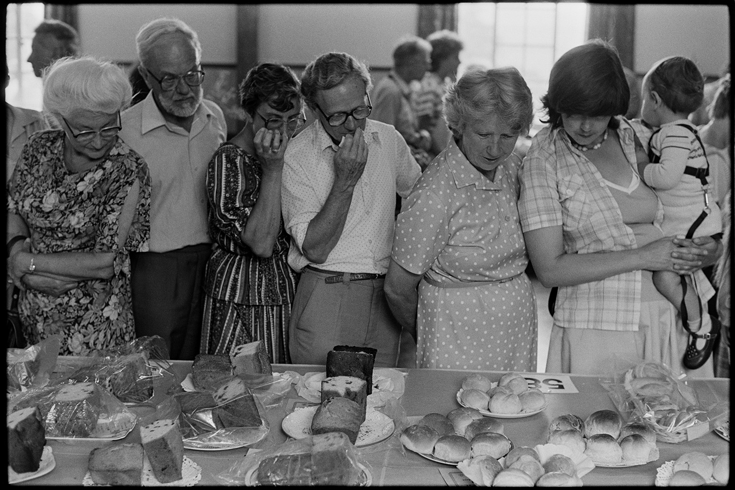 Flower show, people looking at exhibits, women looking at cake, paintings on display.
[Men, women and a child looking at a display of exhibits of fruit cake and bread rolls at Dolton Flower Show in Dolton Village Hall.]