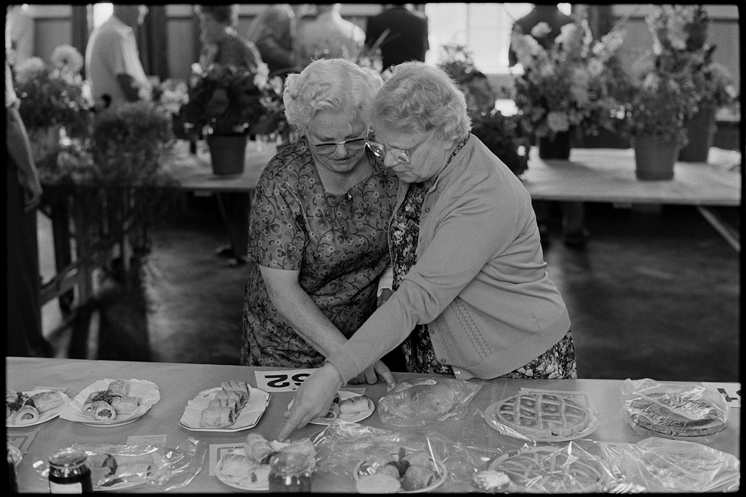 Flower show, people looking at exhibits, women looking at cake, paintings on display.
[Two women looking at exhibits of sausage rolls and flans or tarts on a table at Dolton Flower Show in Dolton Village Hall. A table of floral exhibits is visible in the background.]