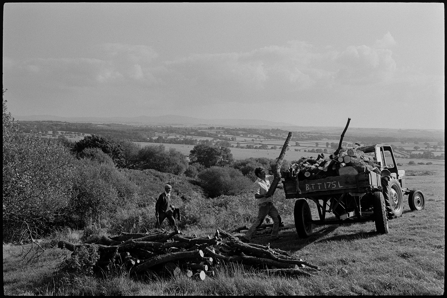 Men cutting trees on moor, and loading logs onto trailer. View to Dartmoor.
[Men loading cut branches into a trailer on Hatherleigh moor, with a landscape  of field and trees in the background. Dartmoor is visible on the horizon.]