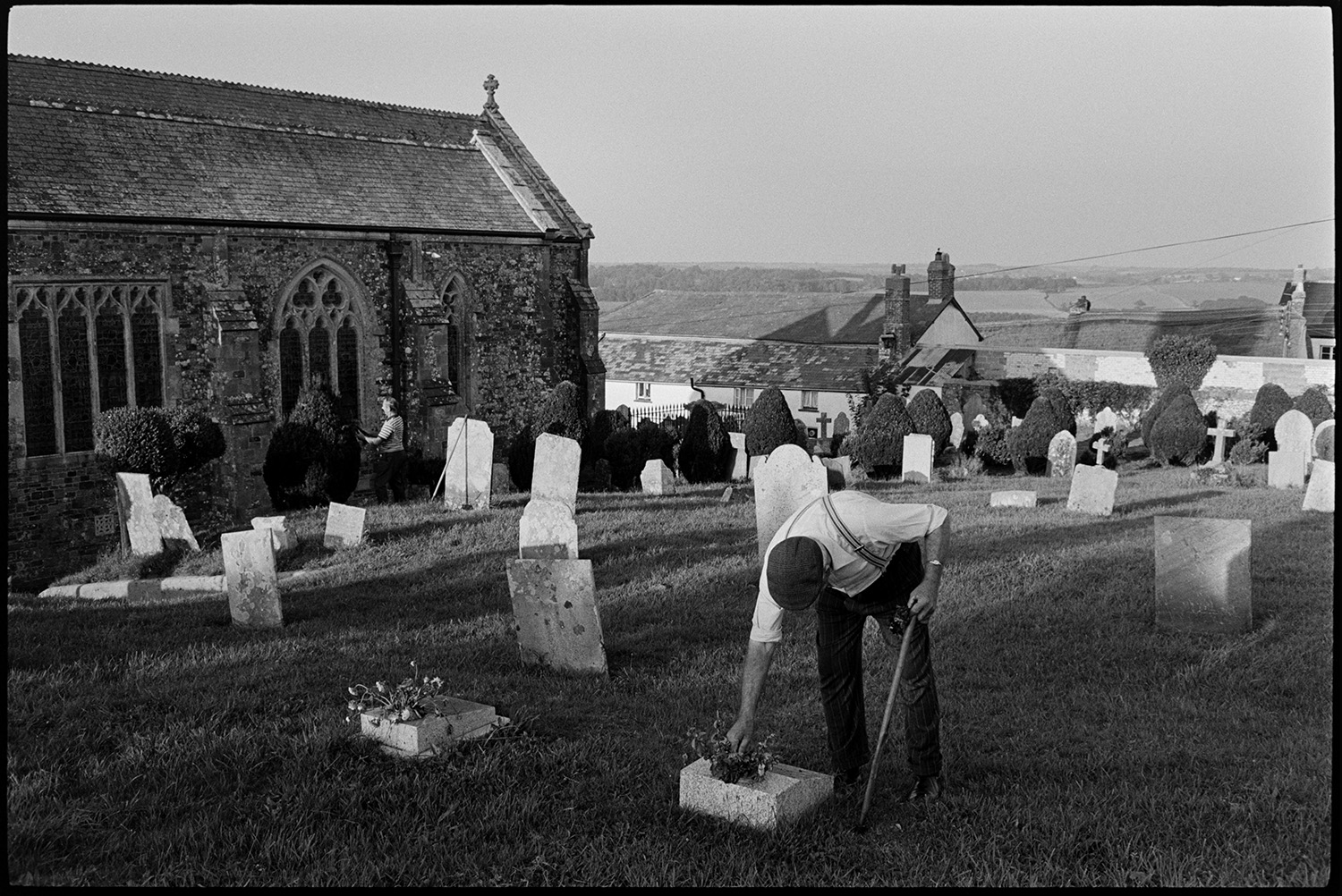 Men working in graveyard, carrying flowers, clipping hedge.
[Men working in Petrockstowe churchyard, tidying gravestones, flowers and clipping hedging. Part of the church is visible and the roofs of village houses can be seen in the background.]