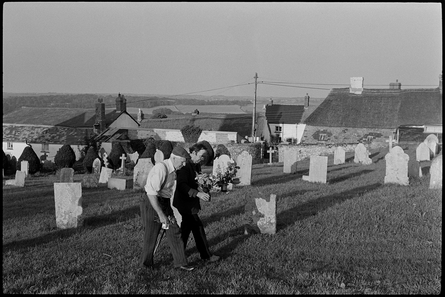 Men working in graveyard, carrying flowers, clipping hedge.
[Two men walking through Petrockstowe churchyard carrying floral displays. Gravestones and clipped bushes are visible in the graveyard and house roofs can be seen in the background.]