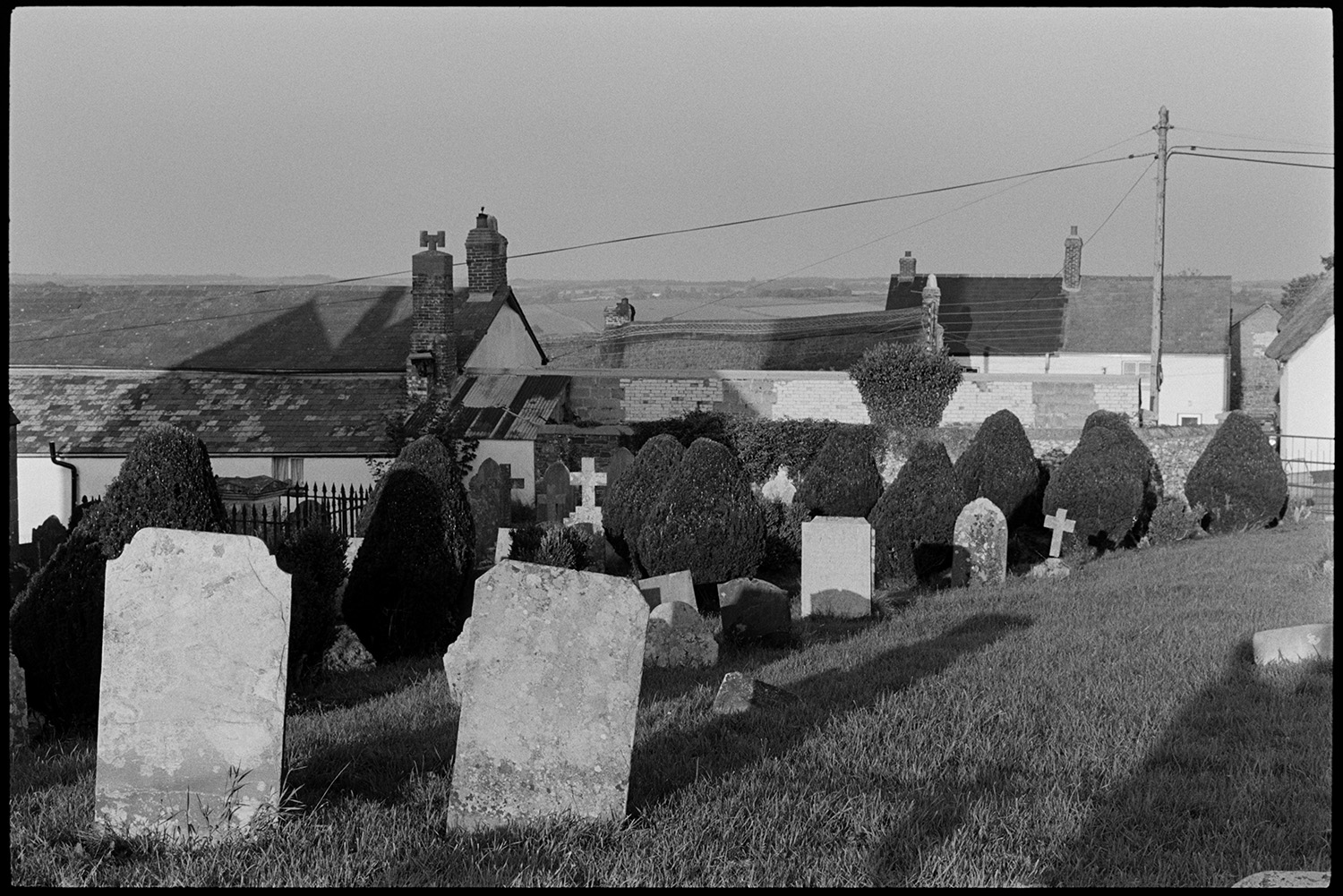Men working in graveyard, carrying flowers, clipping hedge.
[Petrockstowe churchyard with gravestones and clipped yew bushes. Long shadows are stretching across the churchyard and village houses can be seen in the background.]