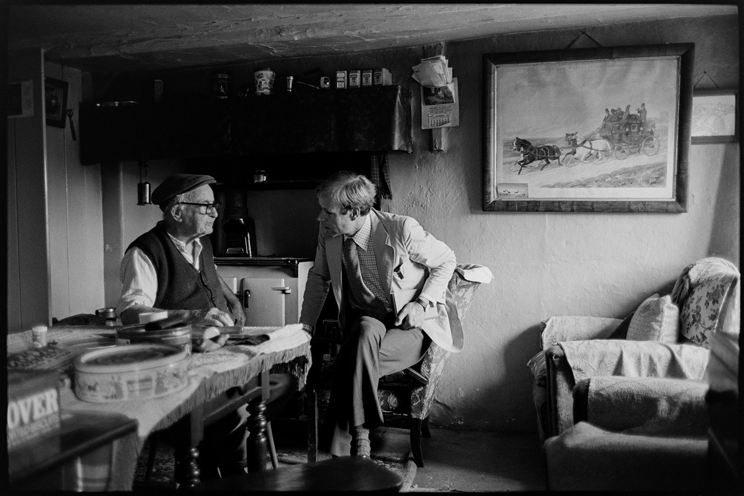 Doctor talking to elderly man patient in his sitting room about going into a home.
[Doctor Richard Westcott talking to Jack Buckingham at Bottreaux Mill, Molland, about going into a care home. The room has a Rayburn, table, armchairs and a large picture hung on the wall of a horse and carriage.]