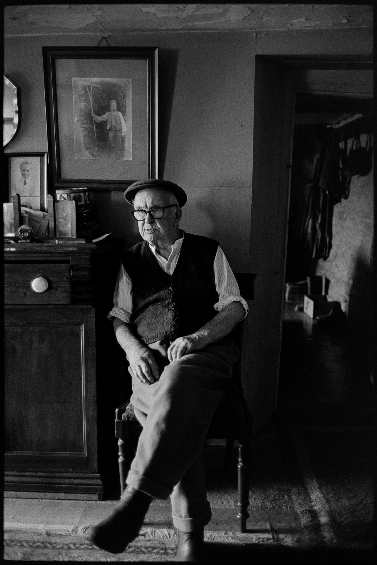 Elderly man seated, talking to doctor, beside cabinet which he made, mirror and photographs.
[Jack Buckingham at Bottreaux Mill, Molland, sitting by a wooden cabinet he had made, with photographs displayed on it. A large family photograph hangs on the wall behind him.]