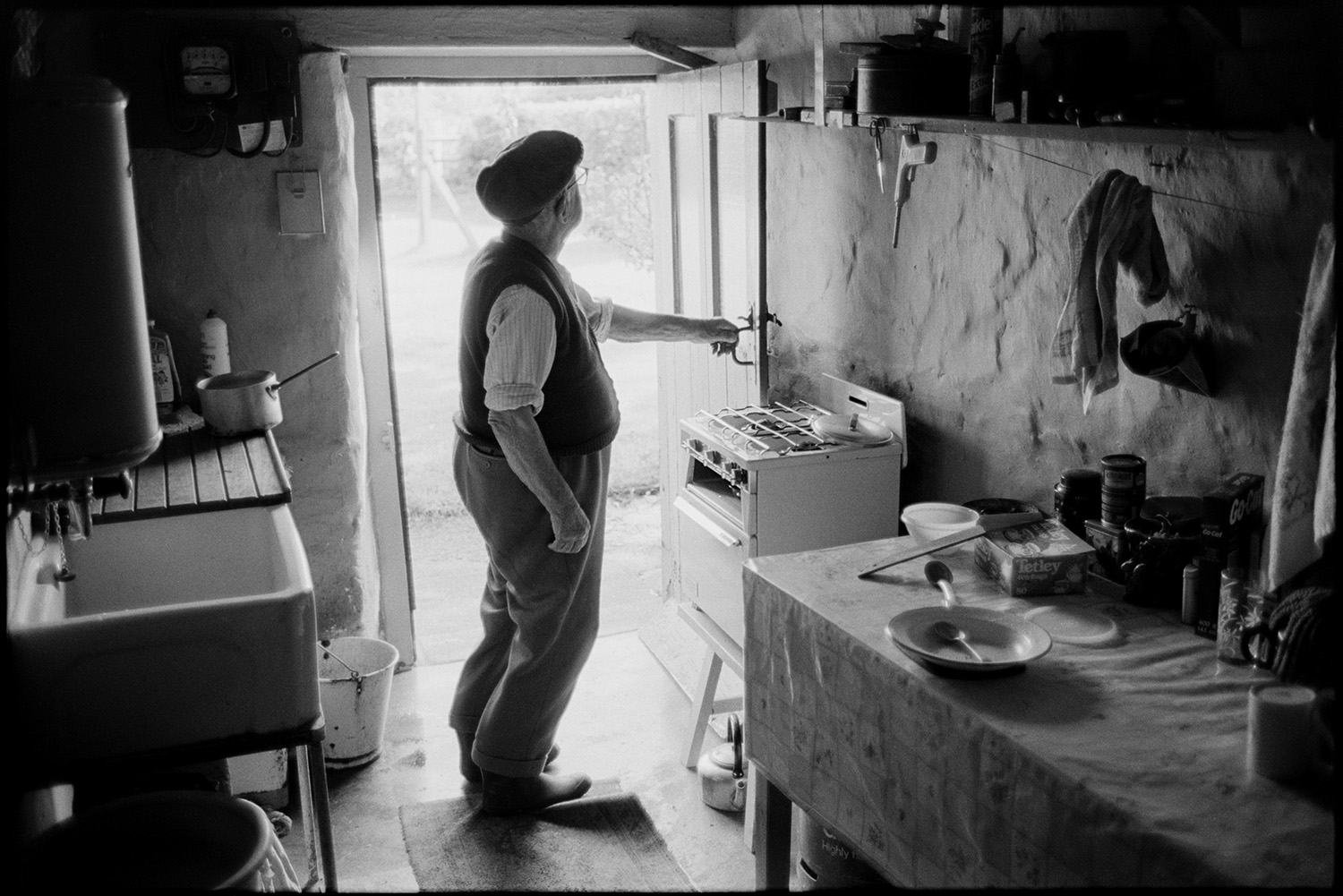 Elderly man standing in his kitchen, side table sink and gas cooker.
[Jack Buckingham at Bottreaux Mill, Molland, standing in his kitchen with a small table, gas cooker, buckets, sink and water heater. He is looking through an open door to the garden beyond.]