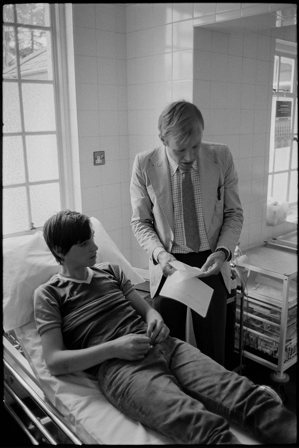 Doctor visiting patients in cottage hospital. Some in bed.
[Doctor Richard Westcott talking to a patient, a young man lying in a bed, in South Molton Cottage Hospital.]
