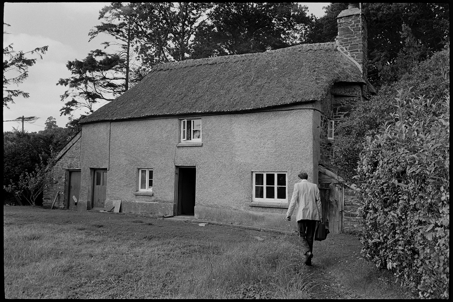 Doctor visiting patient in thatched cottage, walking up to house with bag.
[Doctor Richard Westcott walking up a path to visit a patient in a thatched cottage at Bottreaux Mill, Molland.]