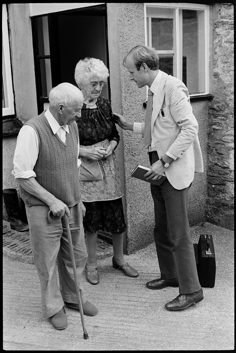 Doctor talking to elderly man and woman on his rounds.
[Doctor Richard Westcott talking to a man and woman outside their home while on his rounds. His case is on the ground behind him and he is holding a notebook.]