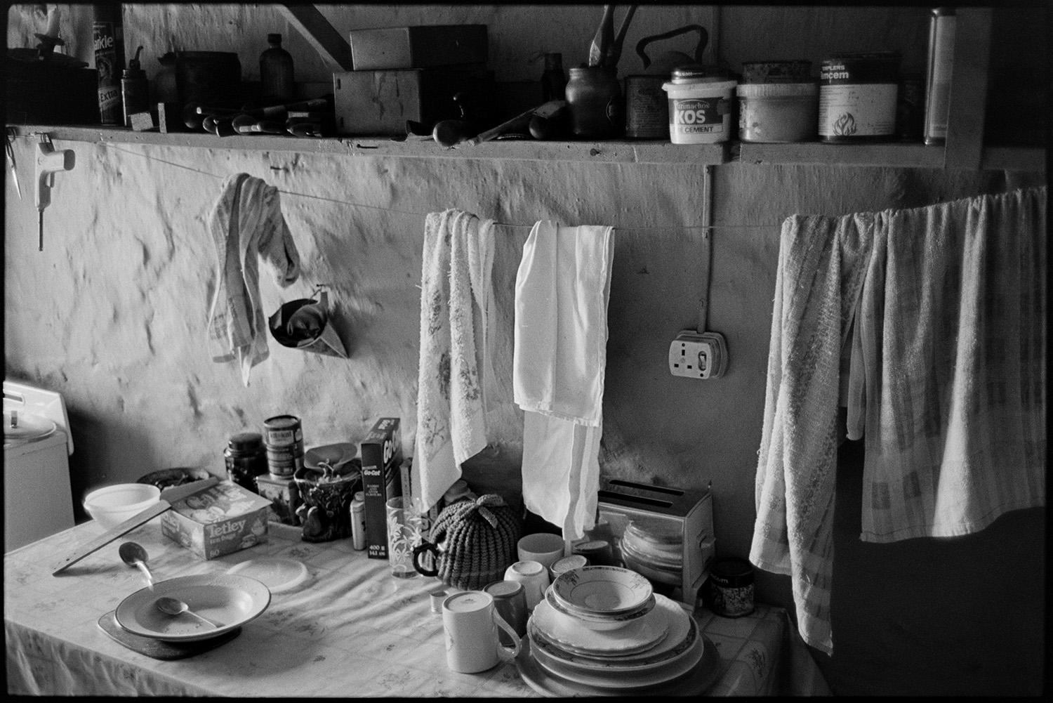 Kitchen sideboard with plates, toaster, teapot and towels.
[The kitchen at Bottreaux Mill, Molland. A table with a toaster, crockery, teapot and groceries on it is visible. Tea towels are hung up to dry over the table and a shelf with various containers and bottles is above the table.]