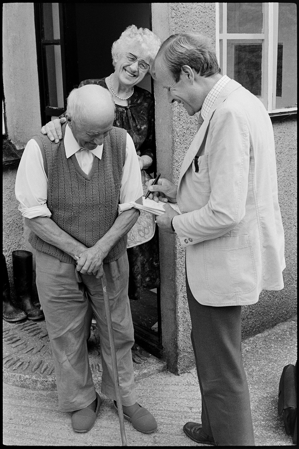 Doctor talking to elderly man and woman on his rounds.
[Doctor Richard Westcott talking to a man and woman in their doorway while on his rounds. He is writing in his notebook.]
