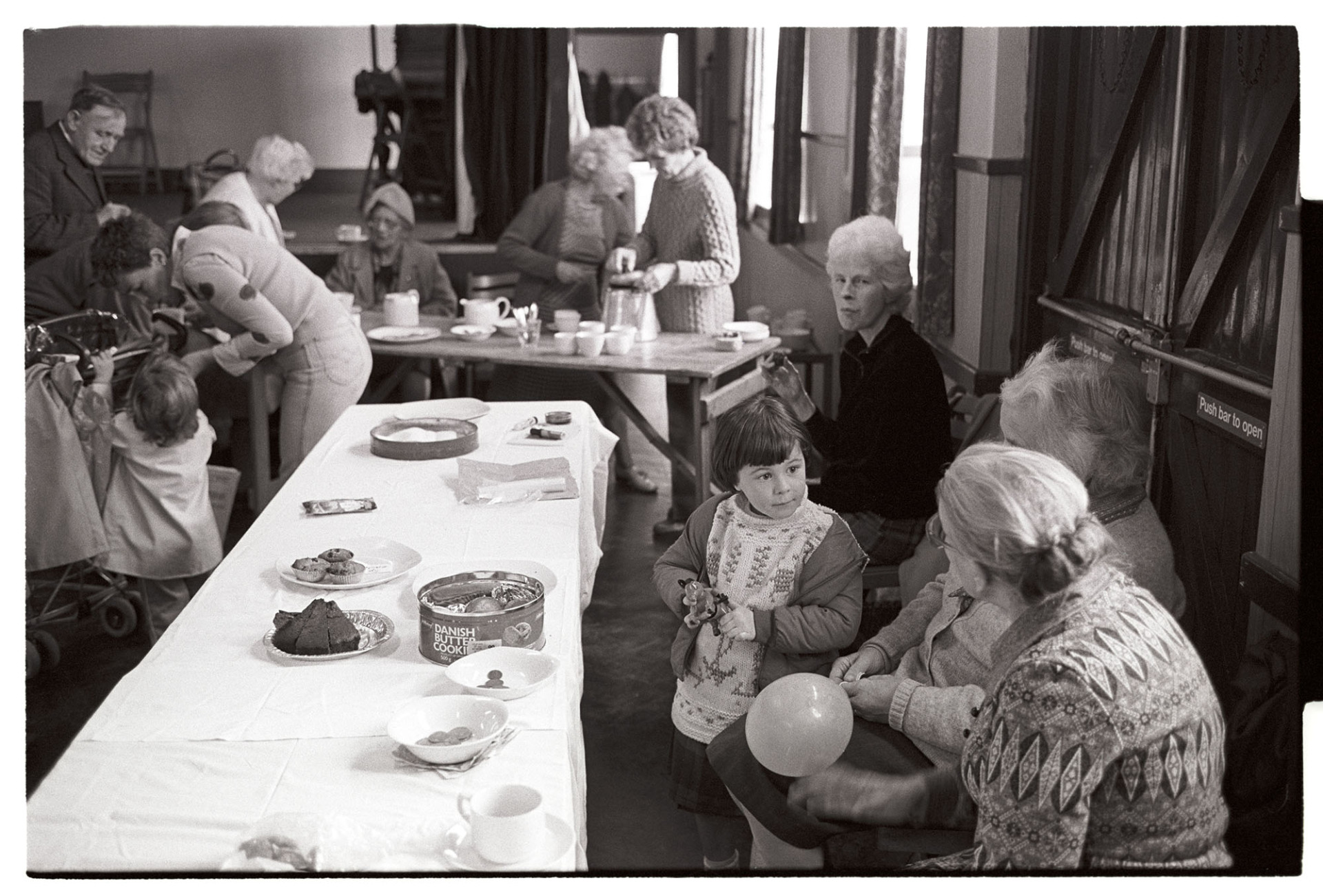 Elderly women and child at playgroup coffee morning. 
[Women and children at a coffee morning at Dolton playgroup in Dolton Village Hall. Two women are serving teas and coffee in the background and a young child, Jenny Agate, is talking to two older women in the foreground. One of the women is holding a balloon and cakes are on the table.]