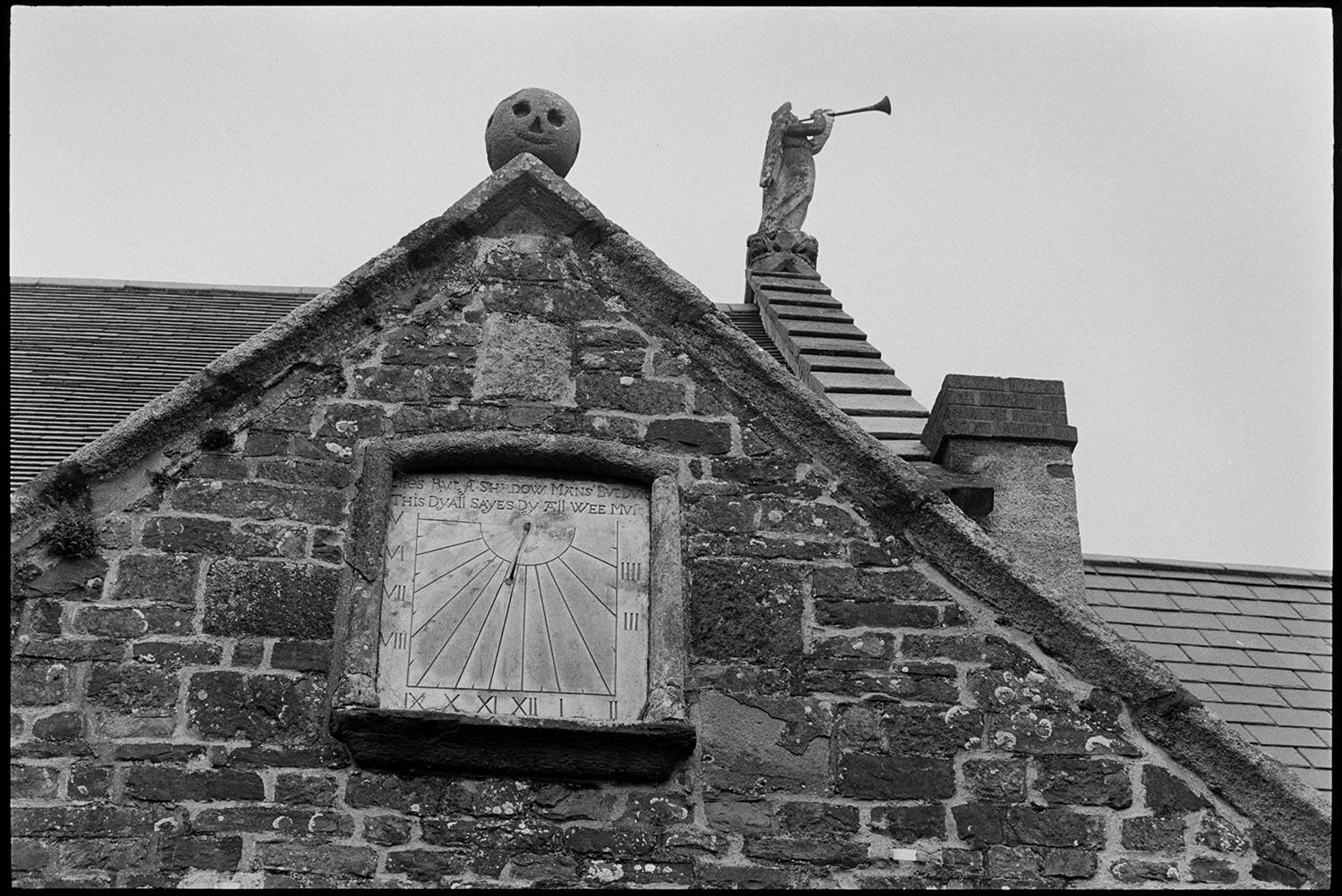 Sundial on front of Victorian railway station.
[A Victorian stone sundial on the front of Kings Nympton railway station. A carved stone face and a stone carving of an angel with a trumpet are also visible on the roof.]