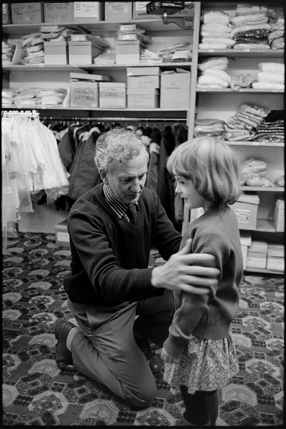 Mother and child buying clothes.
[Ella Ravilious trying on clothes at Singh's clothes shop at South Street, Torrington. A man, possibly the shop manager, is assisting her. Shelves with clothing, shoeboxes and a clothes rails are visible in the background. They are stood on a patterned carpet.]