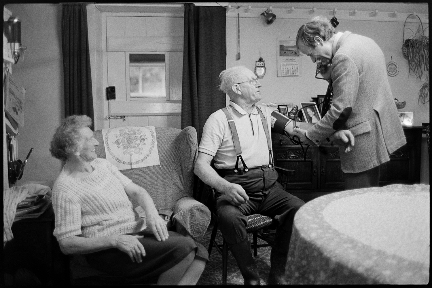 Doctor on his rounds examining men and women patients in their homes.
[Doctor Richard Westcott taking the blood pressure of a man in his house in South Molton. They are in the living room and his wife is sat next to him in an armchair. A table, chairs and sideboard with photographs can be seen in the background, with a wooden door behind a curtain.]