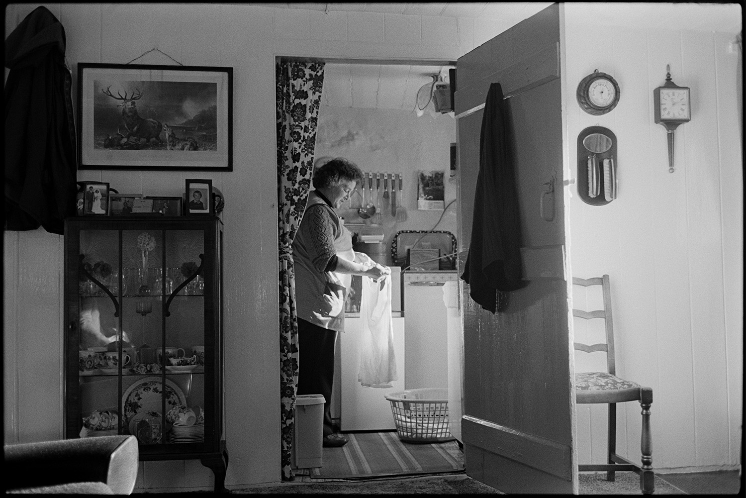 View through sitting room with woman doing washing, washing machine, glass cabinet, picture.
[View through the sitting room to the kitchen at Upcott, Dolton, of Mrs Piper doing the washing. A picture, barometer and clock hang on the wall; and a glass cabinet with china can be seen in the living room. A fridge, washing basket and cooking utensils are visible in the kitchen.]